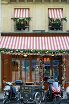A storefront with wooden doors and large windows is adorned with decorative red and white striped awnings. Greenery and festive ornaments, including gold balls, decorate the windows and entrance. Three motorcycles and a cargo bike are parked in front of the establishment. The signage includes calligraphy in gold script and the address of the location.