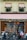 A storefront with wooden doors and large windows is adorned with decorative red and white striped awnings. Greenery and festive ornaments, including gold balls, decorate the windows and entrance. Three motorcycles and a cargo bike are parked in front of the establishment. The signage includes calligraphy in gold script and the address of the location.