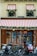 A storefront with wooden doors and large windows is adorned with decorative red and white striped awnings. Greenery and festive ornaments, including gold balls, decorate the windows and entrance. Three motorcycles and a cargo bike are parked in front of the establishment. The signage includes calligraphy in gold script and the address of the location.
