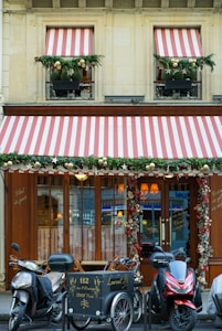 A storefront with wooden doors and large windows is adorned with decorative red and white striped awnings. Greenery and festive ornaments, including gold balls, decorate the windows and entrance. Three motorcycles and a cargo bike are parked in front of the establishment. The signage includes calligraphy in gold script and the address of the location.
