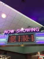 A neon sign with the words 'NOW SHOWING' is prominently displayed above a digital board listing movie showtimes. The board features movies such as Star Wars, Bombshell, and Jumanji 3, along with their respective times. The overall setting suggests a movie theater lobby with ambient neon lighting.