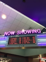 A neon sign with the words 'NOW SHOWING' is prominently displayed above a digital board listing movie showtimes. The board features movies such as Star Wars, Bombshell, and Jumanji 3, along with their respective times. The overall setting suggests a movie theater lobby with ambient neon lighting.