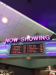 A neon sign with the words 'NOW SHOWING' is prominently displayed above a digital board listing movie showtimes. The board features movies such as Star Wars, Bombshell, and Jumanji 3, along with their respective times. The overall setting suggests a movie theater lobby with ambient neon lighting.