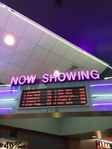 A neon sign with the words 'NOW SHOWING' is prominently displayed above a digital board listing movie showtimes. The board features movies such as Star Wars, Bombshell, and Jumanji 3, along with their respective times. The overall setting suggests a movie theater lobby with ambient neon lighting.