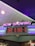 A neon sign with the words 'NOW SHOWING' is prominently displayed above a digital board listing movie showtimes. The board features movies such as Star Wars, Bombshell, and Jumanji 3, along with their respective times. The overall setting suggests a movie theater lobby with ambient neon lighting.