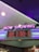 A neon sign with the words 'NOW SHOWING' is prominently displayed above a digital board listing movie showtimes. The board features movies such as Star Wars, Bombshell, and Jumanji 3, along with their respective times. The overall setting suggests a movie theater lobby with ambient neon lighting.