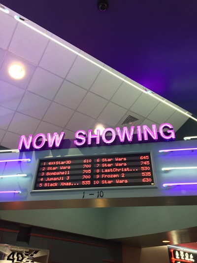 A neon sign with the words 'NOW SHOWING' is prominently displayed above a digital board listing movie showtimes. The board features movies such as Star Wars, Bombshell, and Jumanji 3, along with their respective times. The overall setting suggests a movie theater lobby with ambient neon lighting.
