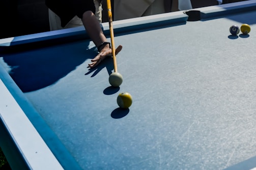 A person is playing billiards on a blue pool table, using a cue to strike a white cue ball positioned near a yellow ball. Shadows cast by the player's arms and cue are visible on the table.