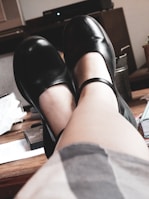 A pair of feet wearing black Mary Jane shoes is elevated and resting on what appears to be a wooden surface in a cozy indoor setting. Various objects such as a metal container, paper, and books or magazines are scattered on the table, suggesting a casual, relaxed environment.