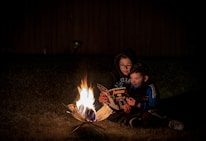 woman wearing eyeglasses sitting near boy reading Avengers comic book beside bonfire during night time