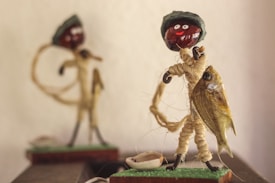 A handcrafted figurine made of twine is holding a fish and wearing a small hat. The face is crafted from a dark round object with two white dots for eyes and a small red painted smile. In the background, there is another similar figure, slightly out of focus.