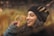 A young woman with long dark curly hair is wearing a black beanie and a denim jacket, holding sunglasses in her hand. The background is a blurred natural setting with earthy tones.