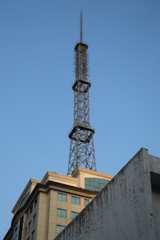 A tall metal communications tower extends upward from the rooftop of a high-rise building. The building has multiple floors with square windows and beige-colored walls. The sky is clear with a bright blue hue.