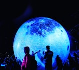 A large, illuminated, blue model of the moon dominates the scene, surrounded by people who are admiring and interacting with it. The surface of the spherical object is textured to resemble the moon's craters and formations, while the surrounding environment is dark, enhancing the glow of the moon's bluish hue. Some individuals are taking photos, and others are pointing or reaching towards the display, creating a sense of wonder and engagement.