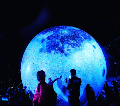 A large, illuminated, blue model of the moon dominates the scene, surrounded by people who are admiring and interacting with it. The surface of the spherical object is textured to resemble the moon's craters and formations, while the surrounding environment is dark, enhancing the glow of the moon's bluish hue. Some individuals are taking photos, and others are pointing or reaching towards the display, creating a sense of wonder and engagement.