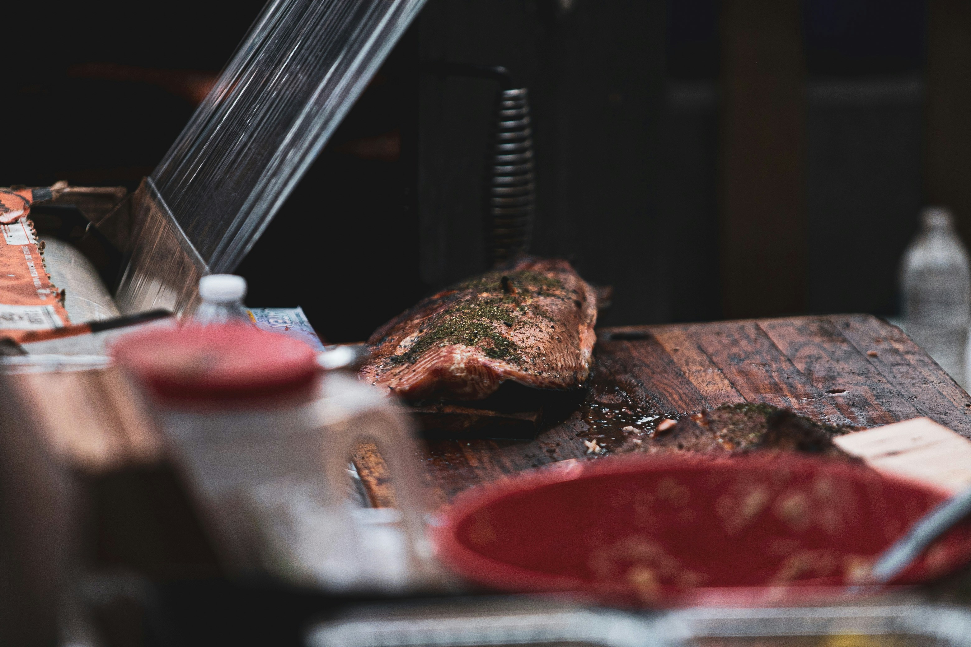 Pedaço de carne temperado sobre tábua de madeira, com utensílios e recipientes ao redor em ambiente de churrasco.