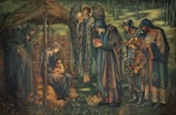 A serene scene depicting several figures in robes around a central seated woman holding a child. An angel with wings stands behind, holding a glowing object. The setting appears to be outdoors with trees and a rustic wooden structure resembling a stable. The figures are engaged in a moment of offering or reverence, with some holding elaborate items, and a crown is placed on the ground.