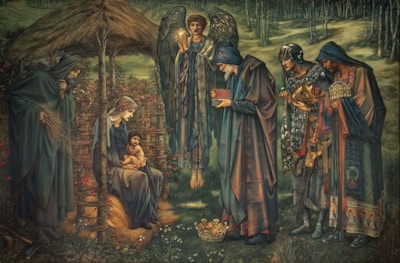 A serene scene depicting several figures in robes around a central seated woman holding a child. An angel with wings stands behind, holding a glowing object. The setting appears to be outdoors with trees and a rustic wooden structure resembling a stable. The figures are engaged in a moment of offering or reverence, with some holding elaborate items, and a crown is placed on the ground.