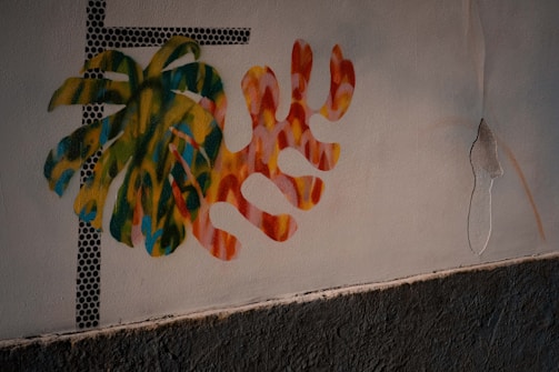 A wall features a colorful mural with abstract plant-like shapes intermingled, painted in vibrant hues of green, yellow, blue, and red. Black dotted lines form a grid pattern on one side, adding a modern touch. The wall is textured at the bottom, with some visible peeling on the side.