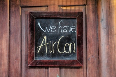 A chalkboard sign is framed in dark wood and fastened to a wooden surface. The chalkboard displays the message 'We have AirCon' written in white and yellow chalk in a casual, handwritten style.