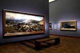 A museum gallery showcases large framed paintings on the walls, featuring a dramatic historical battle scene with a dynamic crowd and battlefield landscape. Soft lighting highlights the artwork, creating a reflective atmosphere.