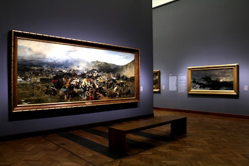 A museum gallery showcases large framed paintings on the walls, featuring a dramatic historical battle scene with a dynamic crowd and battlefield landscape. Soft lighting highlights the artwork, creating a reflective atmosphere.