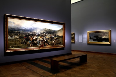 A museum gallery showcases large framed paintings on the walls, featuring a dramatic historical battle scene with a dynamic crowd and battlefield landscape. Soft lighting highlights the artwork, creating a reflective atmosphere.