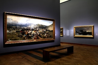 A museum gallery showcases large framed paintings on the walls, featuring a dramatic historical battle scene with a dynamic crowd and battlefield landscape. Soft lighting highlights the artwork, creating a reflective atmosphere.