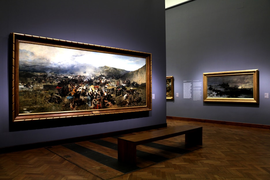 A museum gallery showcases large framed paintings on the walls, featuring a dramatic historical battle scene with a dynamic crowd and battlefield landscape. Soft lighting highlights the artwork, creating a reflective atmosphere.