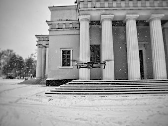 A modern drone hovers in front of a neoclassical building with large columns. The ground and stairs are covered in snow, and snowflakes are visible in the air, creating a wintry atmosphere. The image is in black and white, enhancing the contrast between the drone and the architectural features.