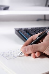 A hand is holding a pen and writing on a notebook page. The paper has a diagram and some handwritten text. A keyboard is visible in the background, slightly out of focus.