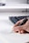 A hand is holding a pen and writing on a notebook page. The paper has a diagram and some handwritten text. A keyboard is visible in the background, slightly out of focus.