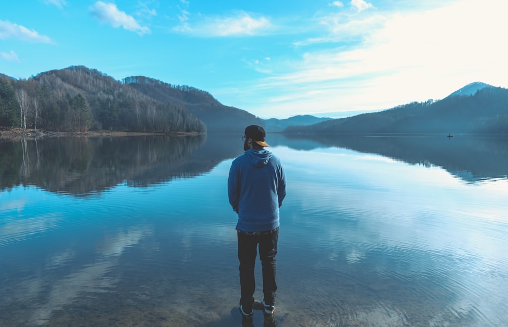 man standing and facing the body of water and mountains during day