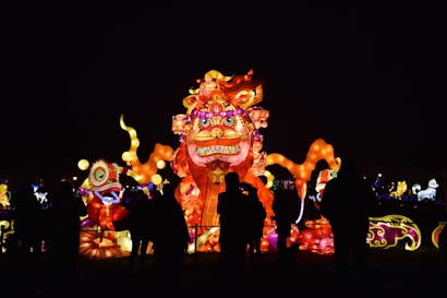 Brightly illuminated lanterns in the shape of fantastical creatures are displayed at night. Silhouettes of people are visible in front of the large, colorful lanterns, which exhibit intricate designs and vibrant colors. The atmosphere appears festive and lively.