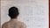 A person is standing in front of a whiteboard that shows flight information. The board is divided into tables presenting domestic and international flights with details such as flight numbers, arrival, and departure data. There are also sections for extra/unscheduled flights. The information is handwritten in blue ink, and the person is facing away from the camera.