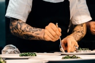 A chef with tattooed arms carefully plating a dish on a white plate, using a spoon. The chef is wearing a white short-sleeved shirt and a black apron, focusing intently on placing the food. The setting appears to be a professional kitchen.