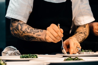 A chef with tattooed arms carefully plating a dish on a white plate, using a spoon. The chef is wearing a white short-sleeved shirt and a black apron, focusing intently on placing the food. The setting appears to be a professional kitchen.