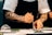 A chef with tattooed arms carefully plating a dish on a white plate, using a spoon. The chef is wearing a white short-sleeved shirt and a black apron, focusing intently on placing the food. The setting appears to be a professional kitchen.