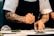 A chef with tattooed arms carefully plating a dish on a white plate, using a spoon. The chef is wearing a white short-sleeved shirt and a black apron, focusing intently on placing the food. The setting appears to be a professional kitchen.