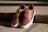 A pair of brown leather sneakers with white soles are placed on concrete steps. The shoes have brown laces and are shown in bright, natural lighting that casts shadows on the textured surface of the steps.