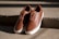 A pair of brown leather sneakers with white soles are placed on concrete steps. The shoes have brown laces and are shown in bright, natural lighting that casts shadows on the textured surface of the steps.