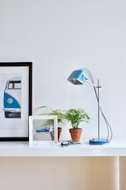 A modern, minimalist desk setup featuring a framed picture of a blue vintage vehicle, a small potted plant, a frame holding a model car, a pair of glasses, and a sleek blue desk lamp. The overall arrangement is placed on a white desk with a light gray background, creating an airy and organized atmosphere.