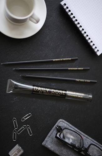 A neatly arranged desk setup includes a white coffee cup, a spiral-bound notebook with a grid pattern, three pencils with engraved text, a large pencil case labeled 'Daily Dose of Inspiration', paper clips, and a pair of eyeglasses inside a soft case. The background is a dark textured surface.