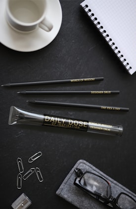 A neatly arranged desk setup includes a white coffee cup, a spiral-bound notebook with a grid pattern, three pencils with engraved text, a large pencil case labeled 'Daily Dose of Inspiration', paper clips, and a pair of eyeglasses inside a soft case. The background is a dark textured surface.