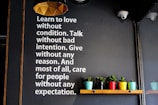 A wall with motivational text encouraging love, unconditional care, and selflessness is displayed in white letters. Below the text, a wooden shelf holds several small potted plants in colorful containers, adding vibrancy to the scene. The lighting fixture above has a geometric, modern design.