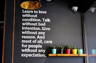 A wall with motivational text encouraging love, unconditional care, and selflessness is displayed in white letters. Below the text, a wooden shelf holds several small potted plants in colorful containers, adding vibrancy to the scene. The lighting fixture above has a geometric, modern design.