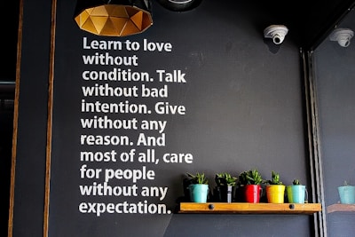 A wall with motivational text encouraging love, unconditional care, and selflessness is displayed in white letters. Below the text, a wooden shelf holds several small potted plants in colorful containers, adding vibrancy to the scene. The lighting fixture above has a geometric, modern design.