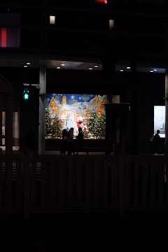 A festive display with Santa Claus is illuminated in the background, surrounded by snowy scenery and Christmas trees adorned with lights. Silhouettes of people pass by in front, with a dimly lit urban setting surrounding the scene.