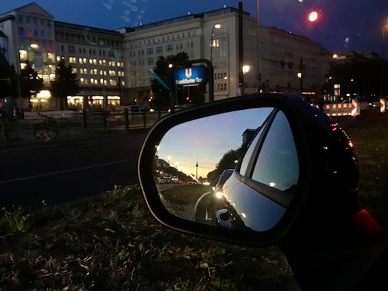 An urban street scene during twilight with a car’s side mirror reflecting a skyline view. Buildings are illuminated with street lights, and a prominent subway sign for 'Frankfurter Tor' is visible. The mirror shows a sunset with colorful hues and a famous tower in the distance.