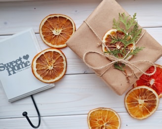 A neatly wrapped gift with brown paper, adorned with dried orange slices and green foliage, is placed on a light wooden surface. Beside the gift lies a book or notebook with the text 'Sweet Home' visible, surrounded by additional dried orange slices.